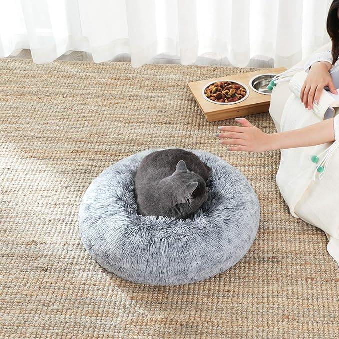 Gray cat curled up in a fluffy round gray pet bed on a woven rug next to a wooden pet food and water bowl